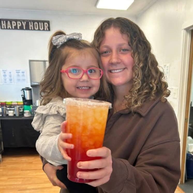 A smiling guest holding a little girl and a sunset-colored loaded tea under Orchard Nutrition's Happy Hour sign